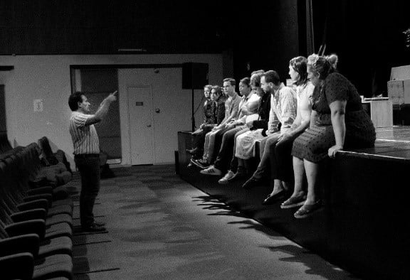 Nicolas Bret-Morel gives instructions to the actors in his theatre company seated on the edge of the stage.